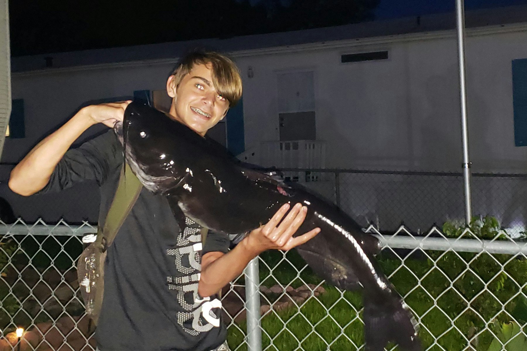 young man holding large catfish with fence and trailer in background