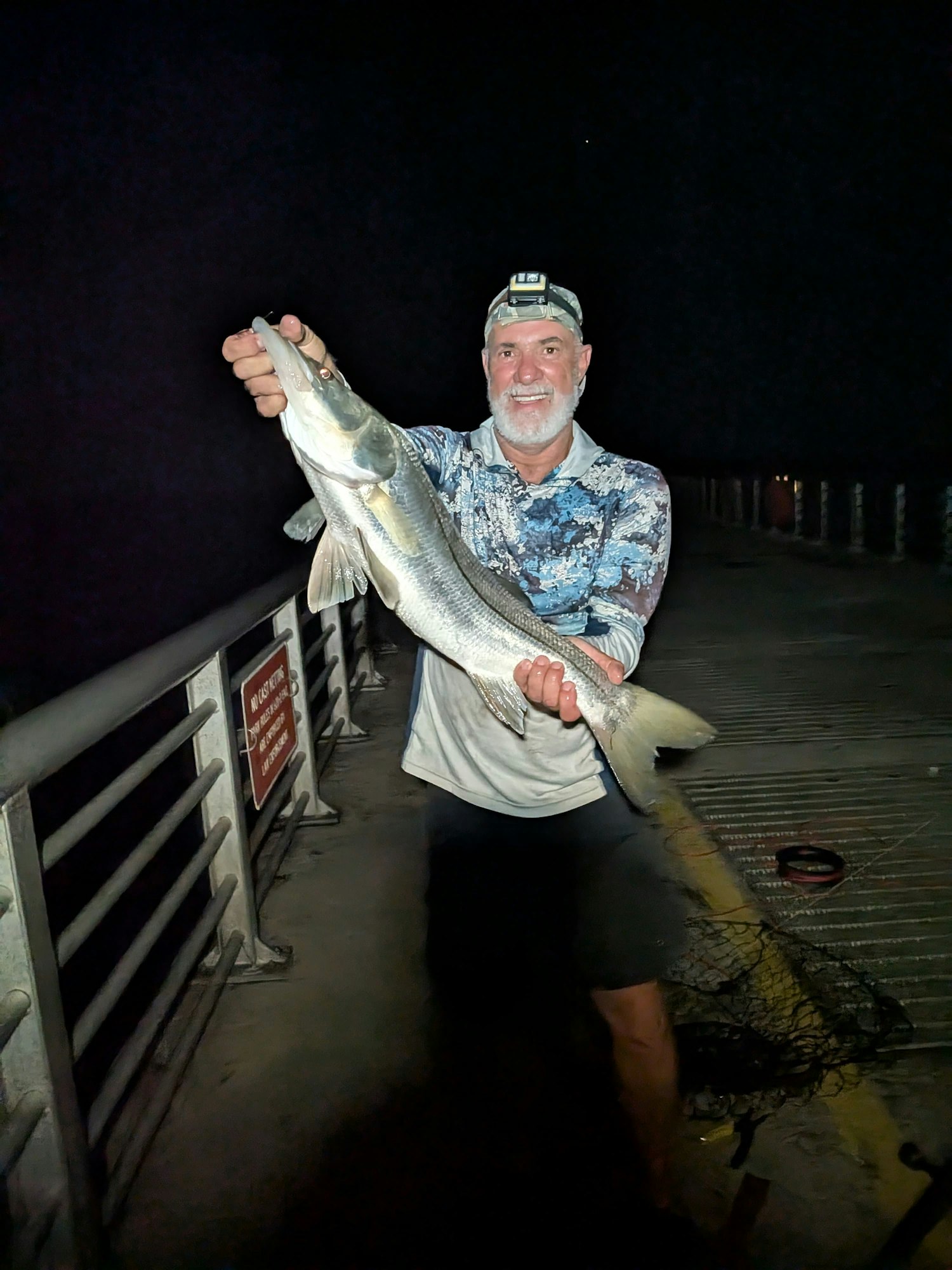 A person with a headlamp holds a large fish on a pier at night.
