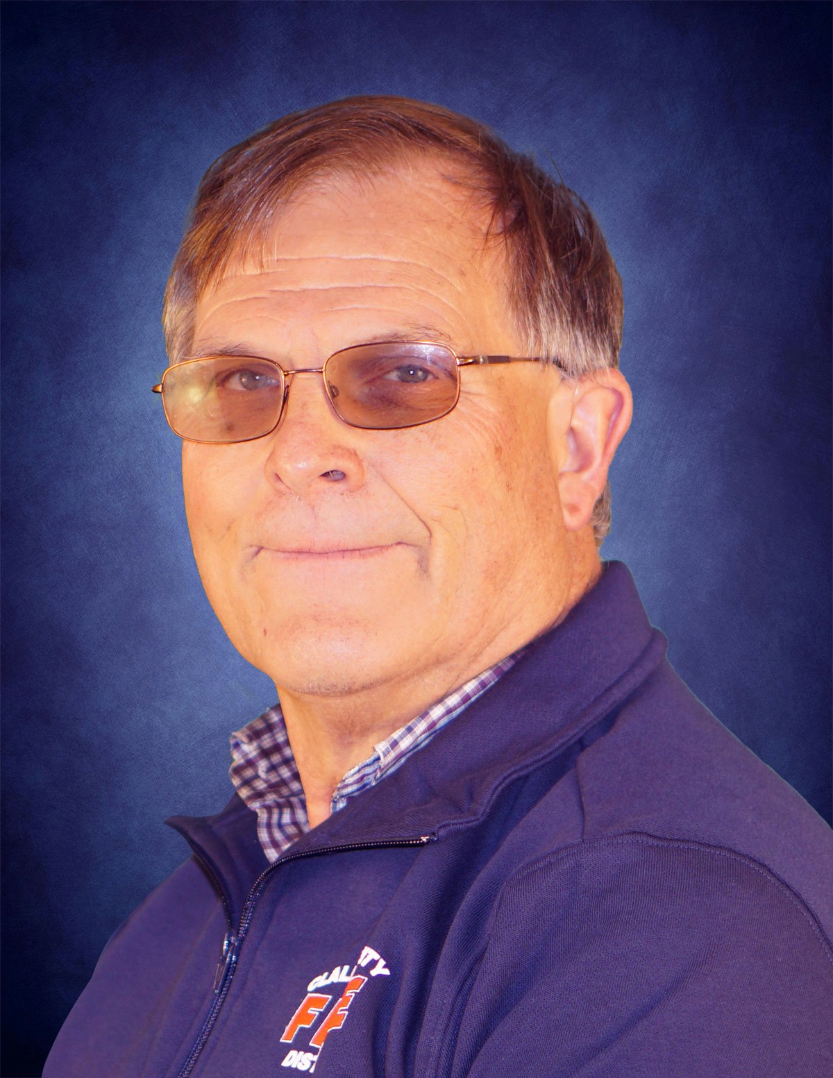 A person wearing glasses, a navy blue jacket with an insignia, and a checkered shirt, looking at the camera against a blue background.