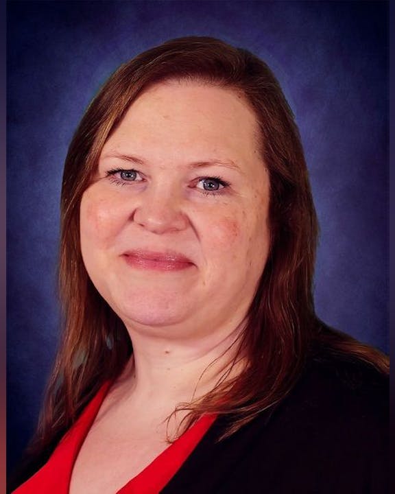 A woman with long brown hair, wearing a red top and dark jacket, against a dark background.