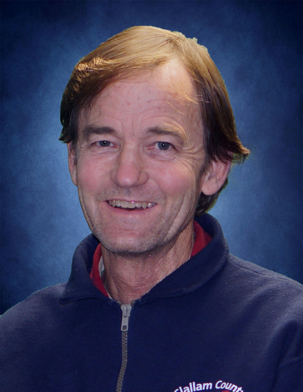 Portrait of a smiling person wearing a dark zip-up jacket against a blue background.