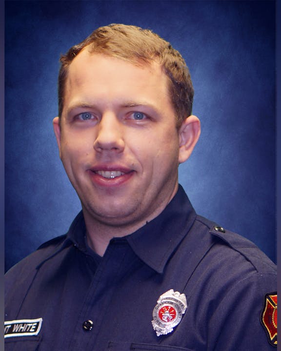 A person in a dark firefighter uniform with badge and name tag, smiling, in front of a blue backdrop.