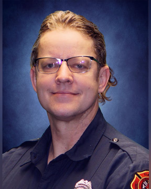 A person wearing glasses and a uniform with a badge on a blue background.