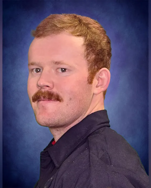 The image features a man with short, wavy hair and a mustache, wearing a dark jacket against a blue background.