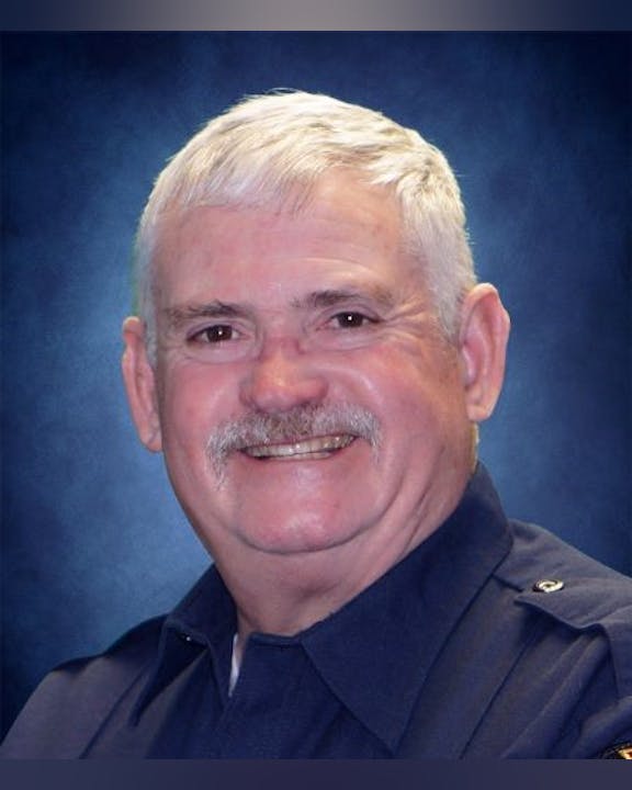 A smiling man with a mustache in a blue uniform against a blue background.