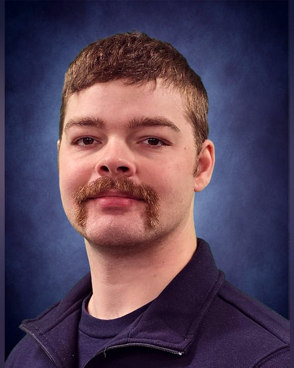 A person with short hair and a mustache against a blue background.