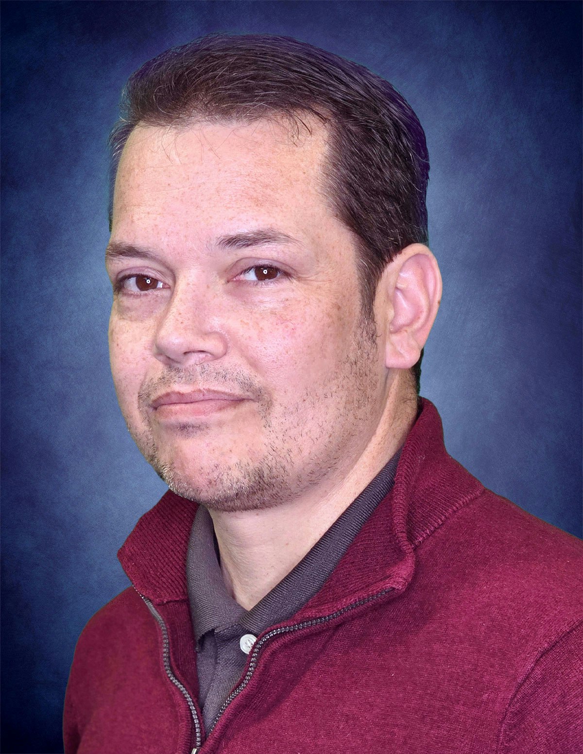 The image shows a man with short hair, wearing a maroon zip-up sweater over a dark shirt, posed against a blue background.