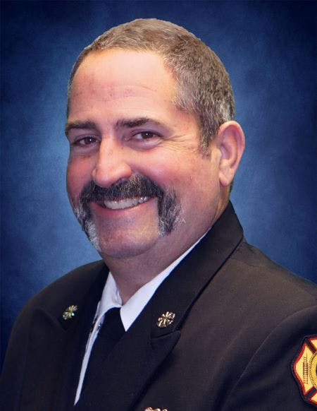 The image shows a smiling man in fire department attire, featuring a distinctive mustache and a friendly expression.