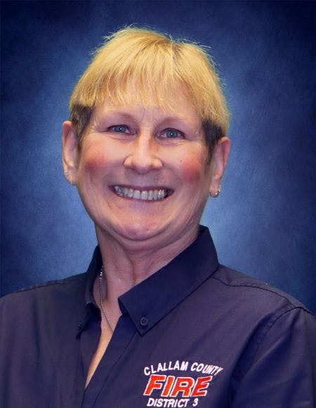 The image shows a woman with short blonde hair, smiling while wearing a dark shirt with "Clallam County Fire District 3" on it.