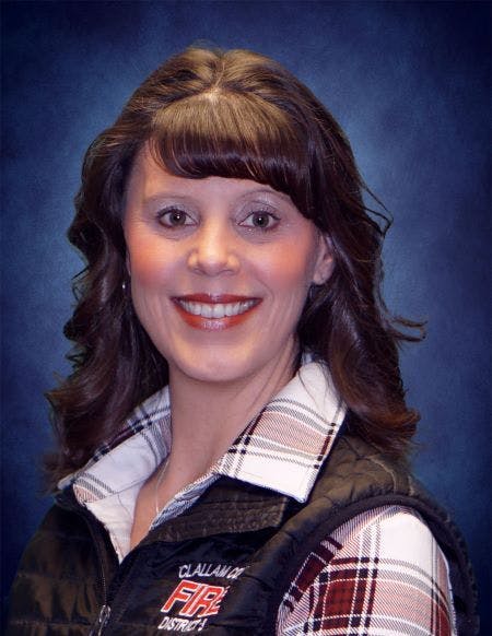 A woman with curly hair smiles in a black vest over a plaid shirt, set against a blue background.