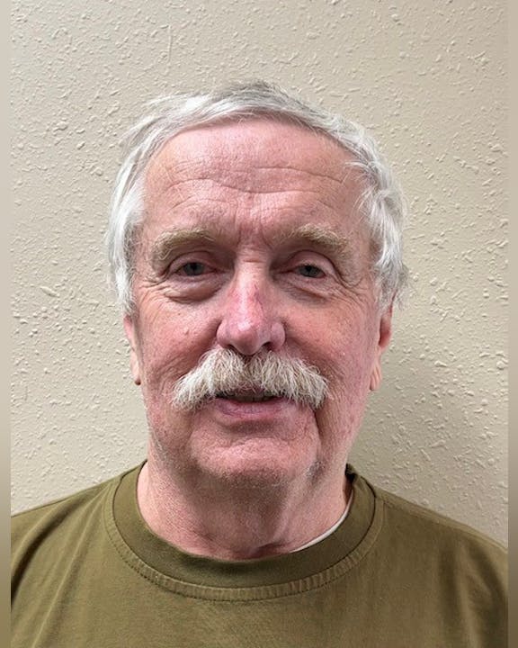 A man with gray hair and a mustache, wearing a brown shirt stands in front of a textured beige wall.