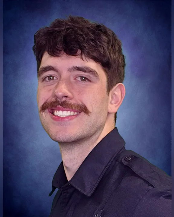 The image shows a smiling young man with dark, curly hair and a mustache, wearing a black shirt against a blue background.