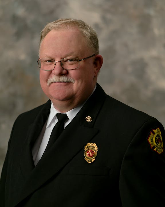 A man in a suit with a badge on his jacket, posing against a neutral background.