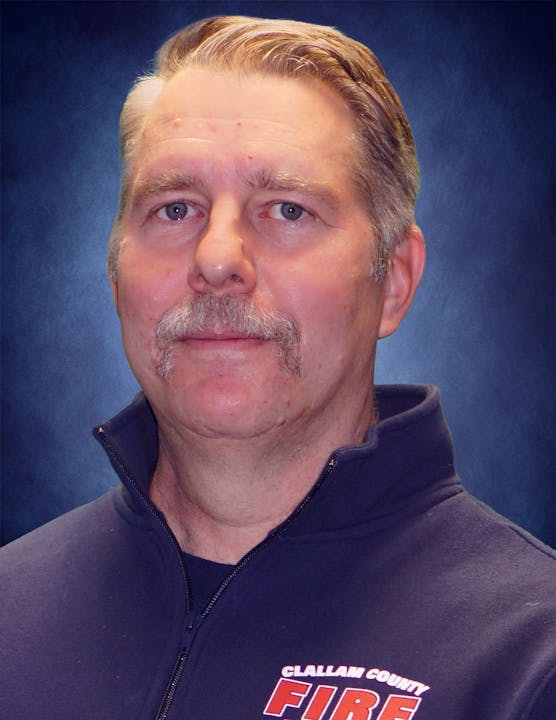 A man with a mustache wearing a "Clallam County FIRE" jacket against a blue background.