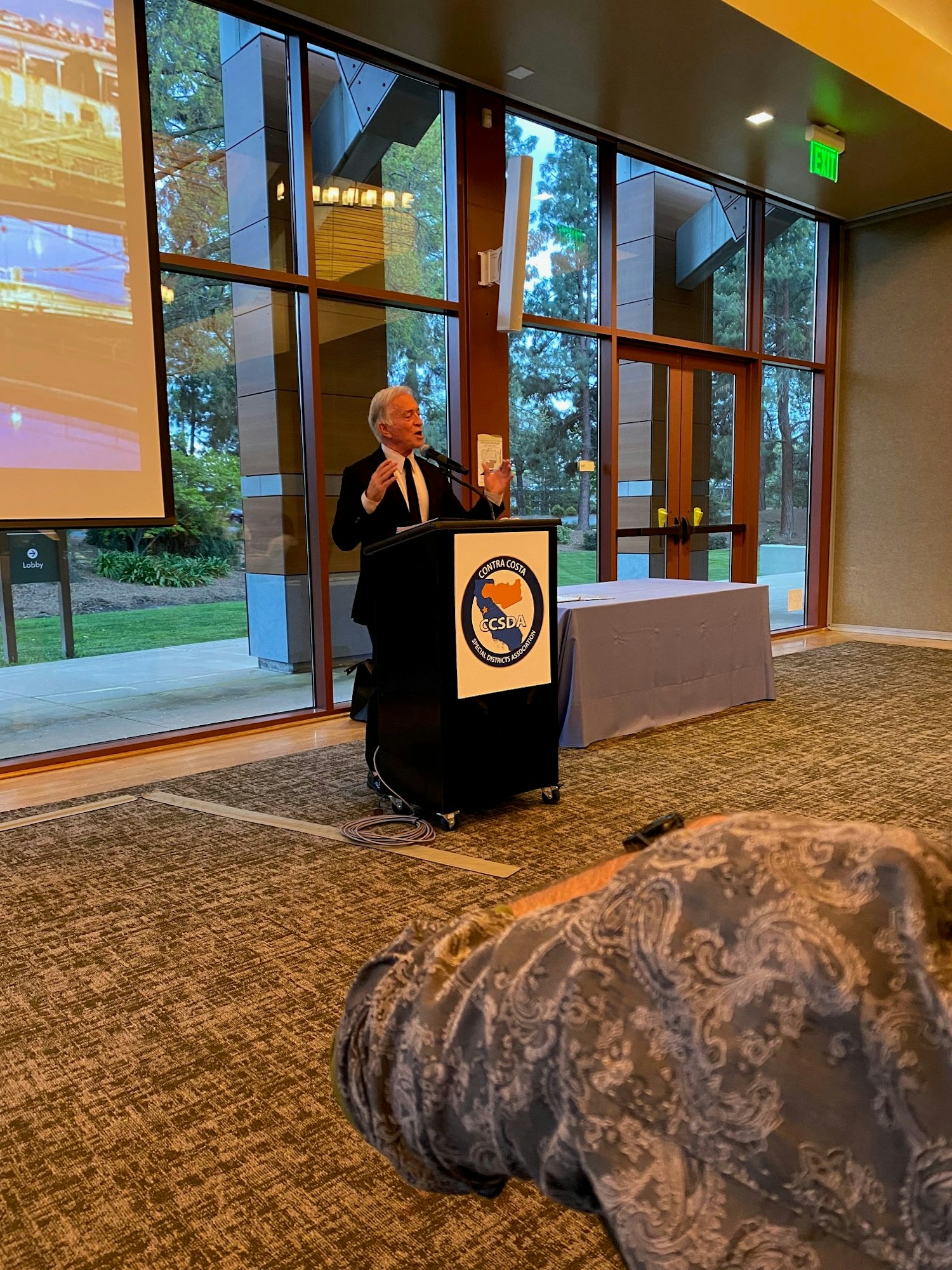 A speaker at a podium with a CCSDA logo, addressing an audience in a conference room with large windows.