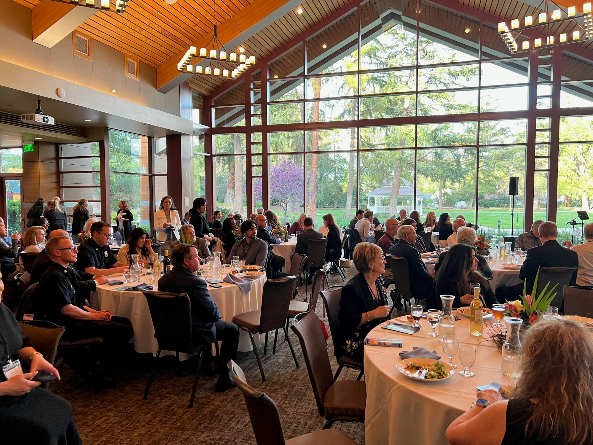 People seated at round tables in a large, windowed banquet hall, attending an event or dinner.