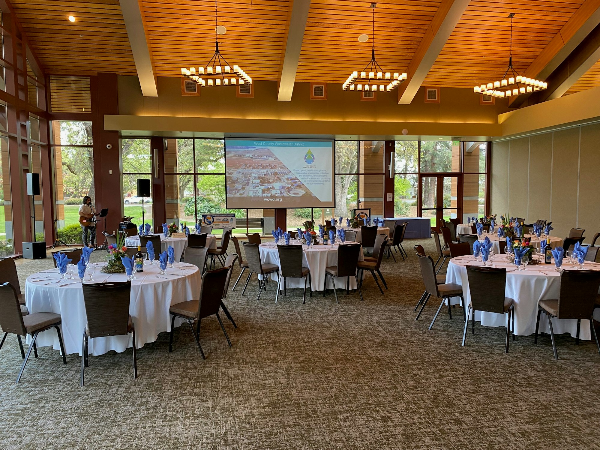 A banquet hall set up for an event with round tables, blue napkins, a projection screen, and a performer with a guitar.