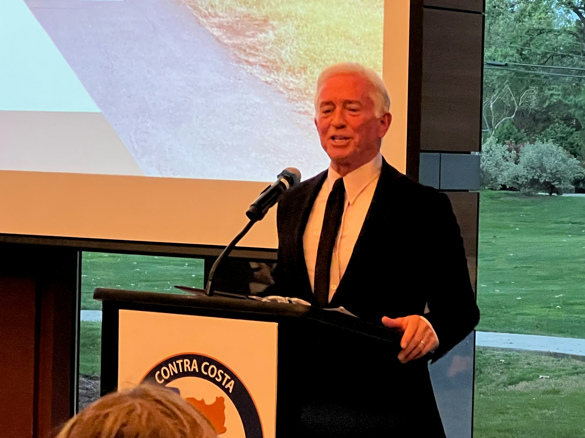 A man in a suit speaking at a podium labeled "Contra Costa," with a microphone and a projected image behind him.