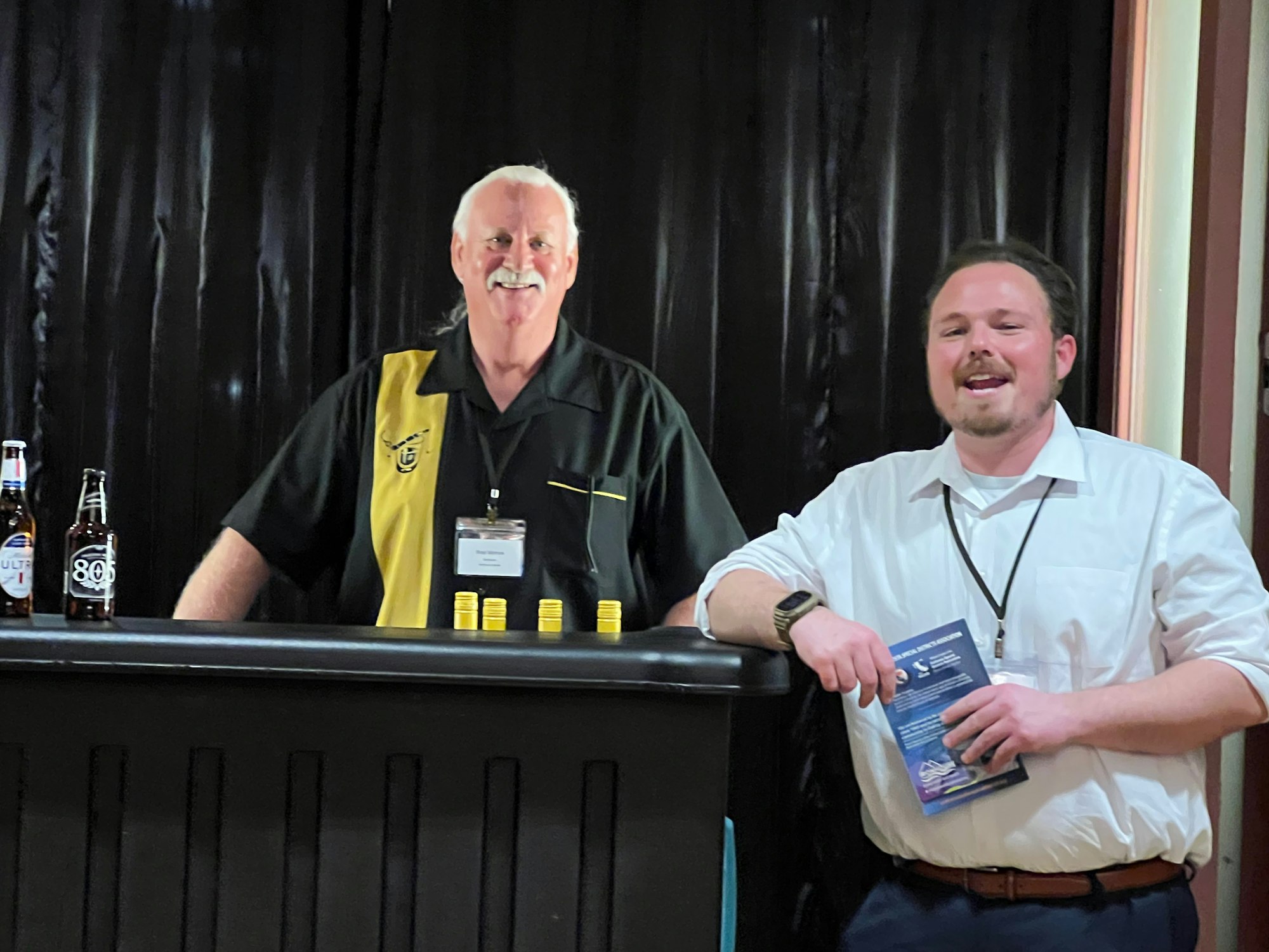 Two men standing at a bar with beer bottles and yellow caps, smiling at the camera.