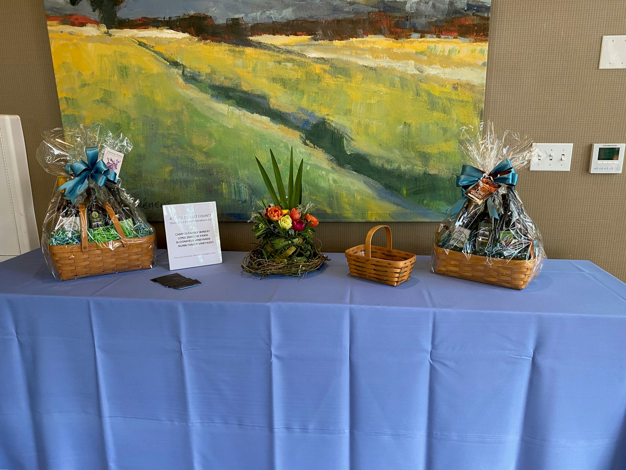 Two gift baskets and a floral arrangement on a table with a blue cloth, under a landscape painting.