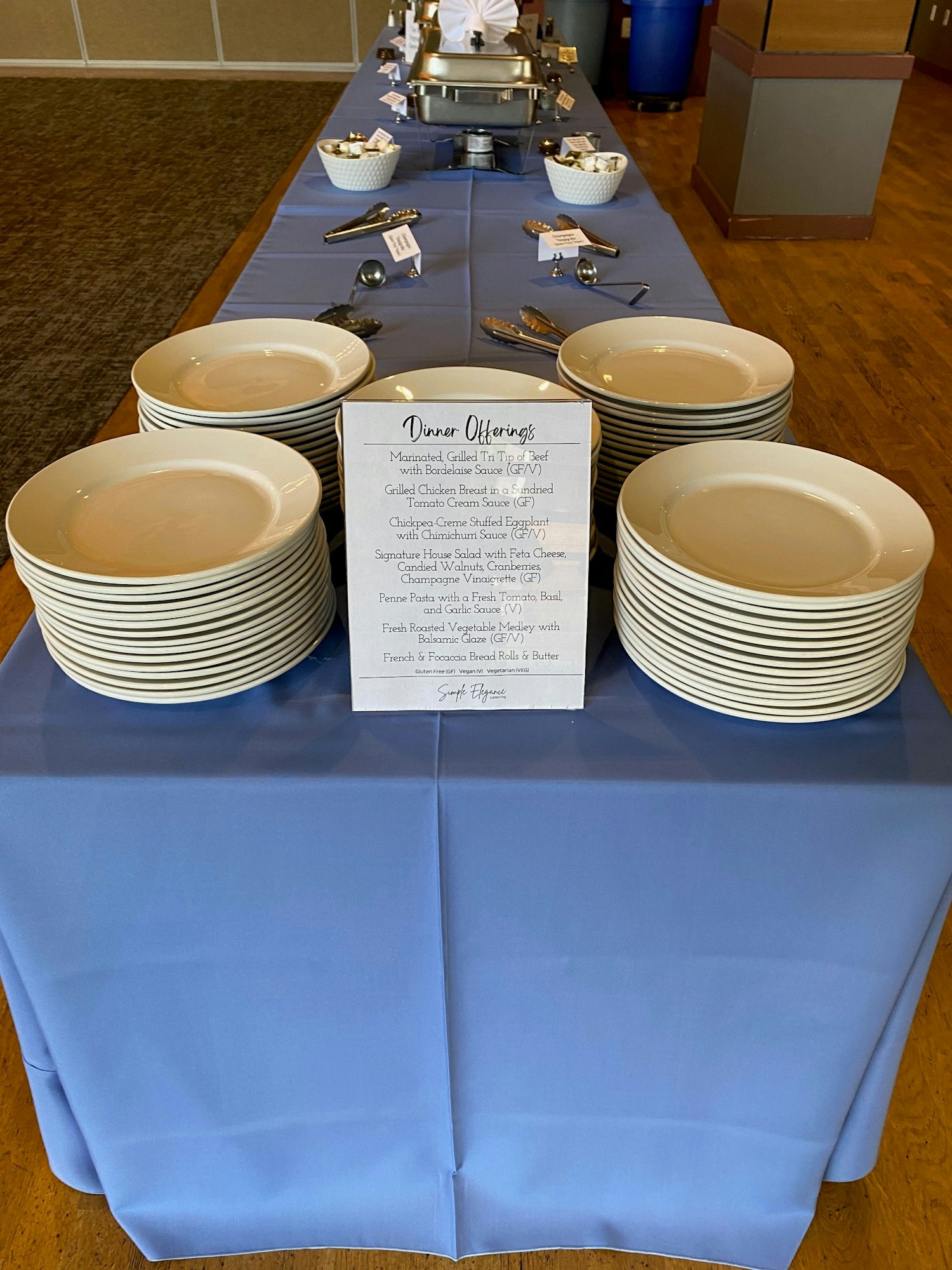 Buffet setup with stacked plates, a menu of dinner offerings, and serving utensils on a blue tablecloth.