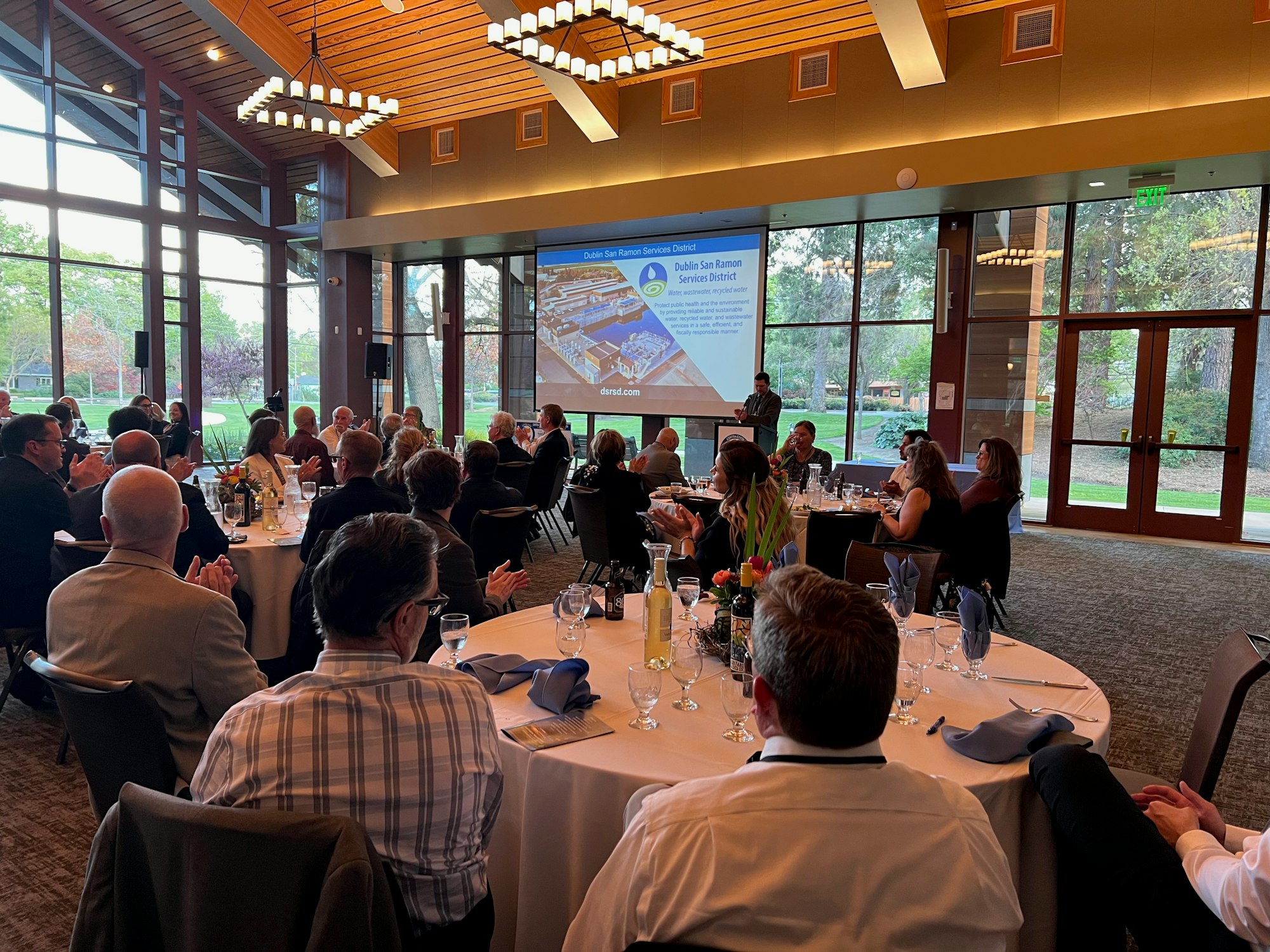 A group of people seated at tables in a hall, listening to a presentation on a screen.
