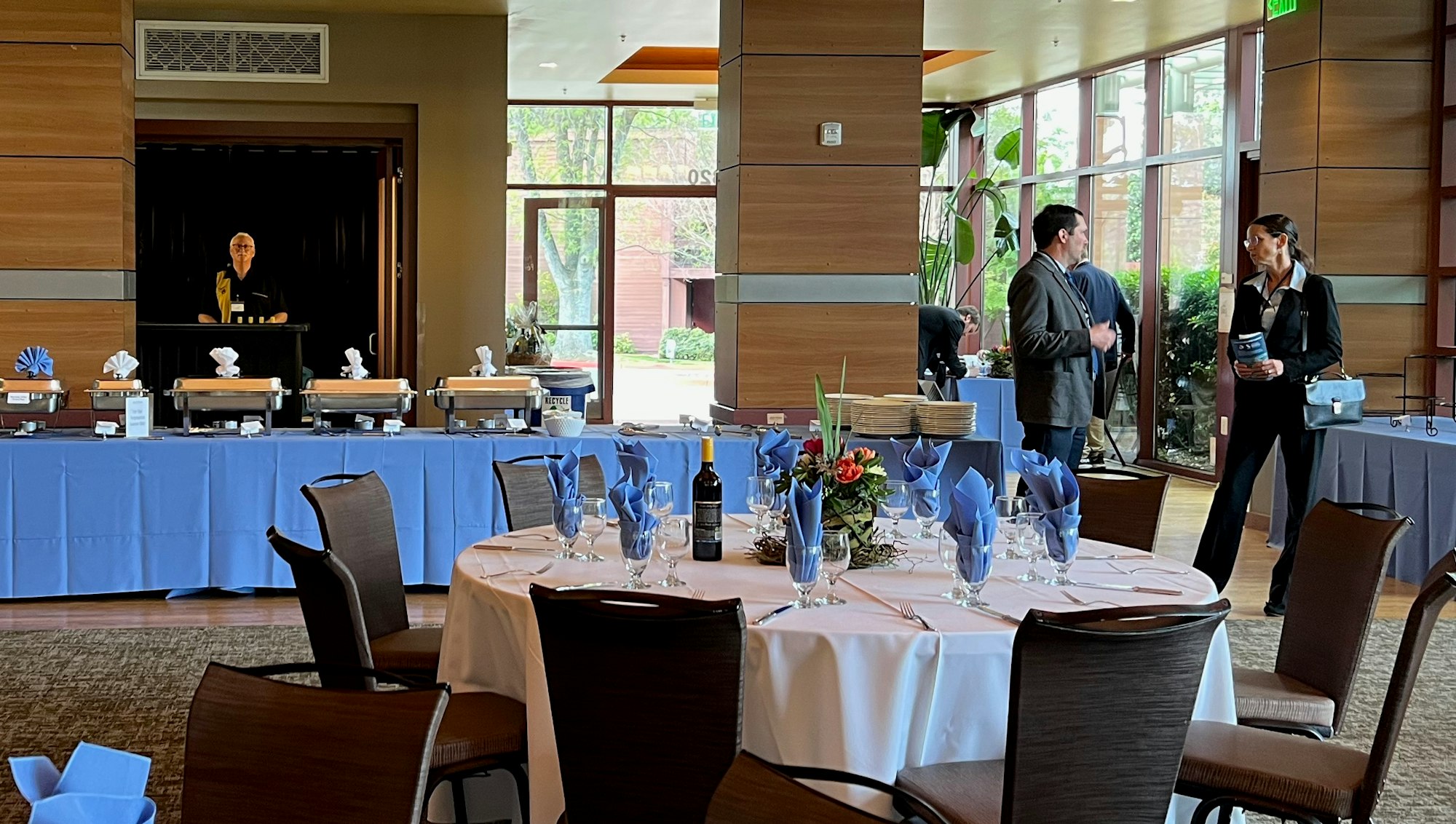 A banquet room with a buffet setup, tables with blue napkins, and two people talking.