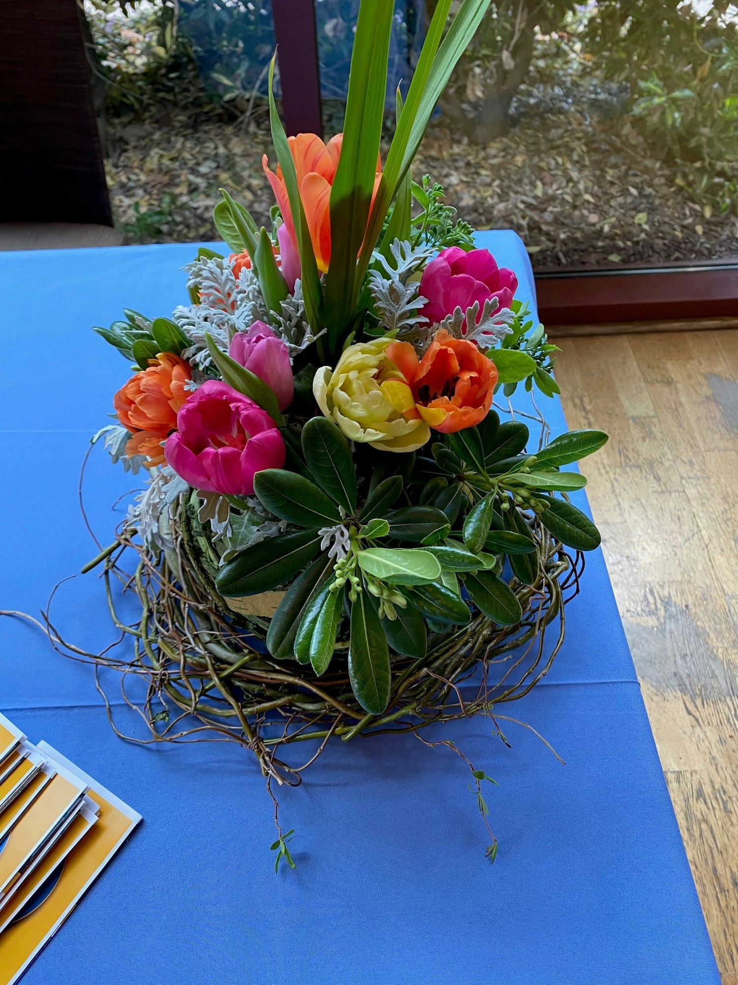 A colorful flower arrangement with pink, orange, and yellow blooms on a blue tablecloth near a window.