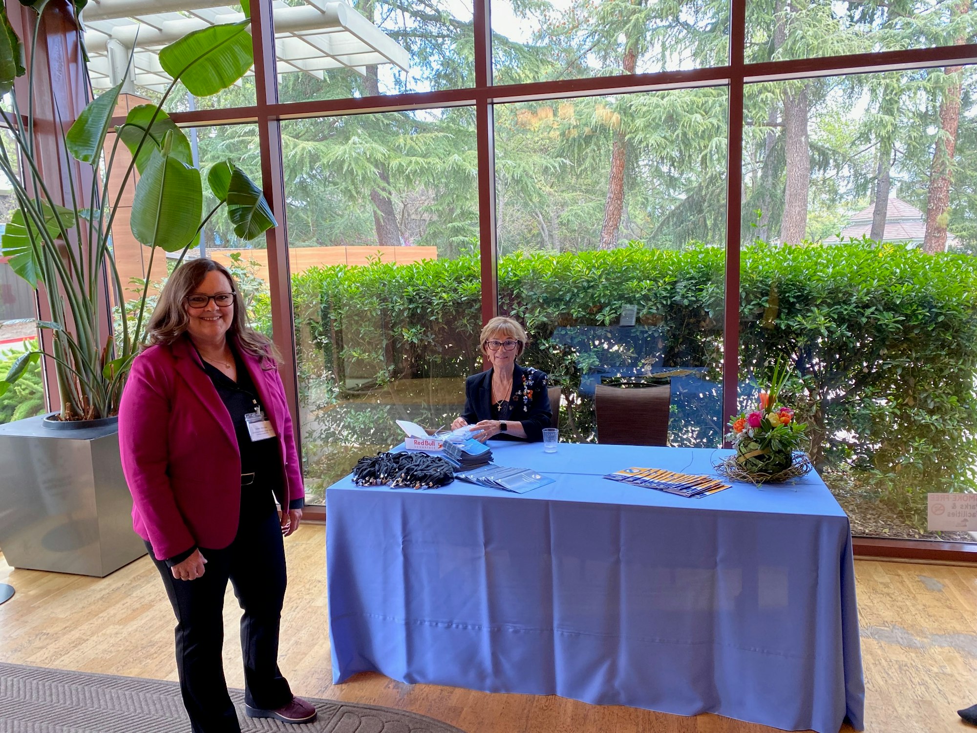 Two people at a registration table with brochures and lanyards, beside a large window with plants outside.
