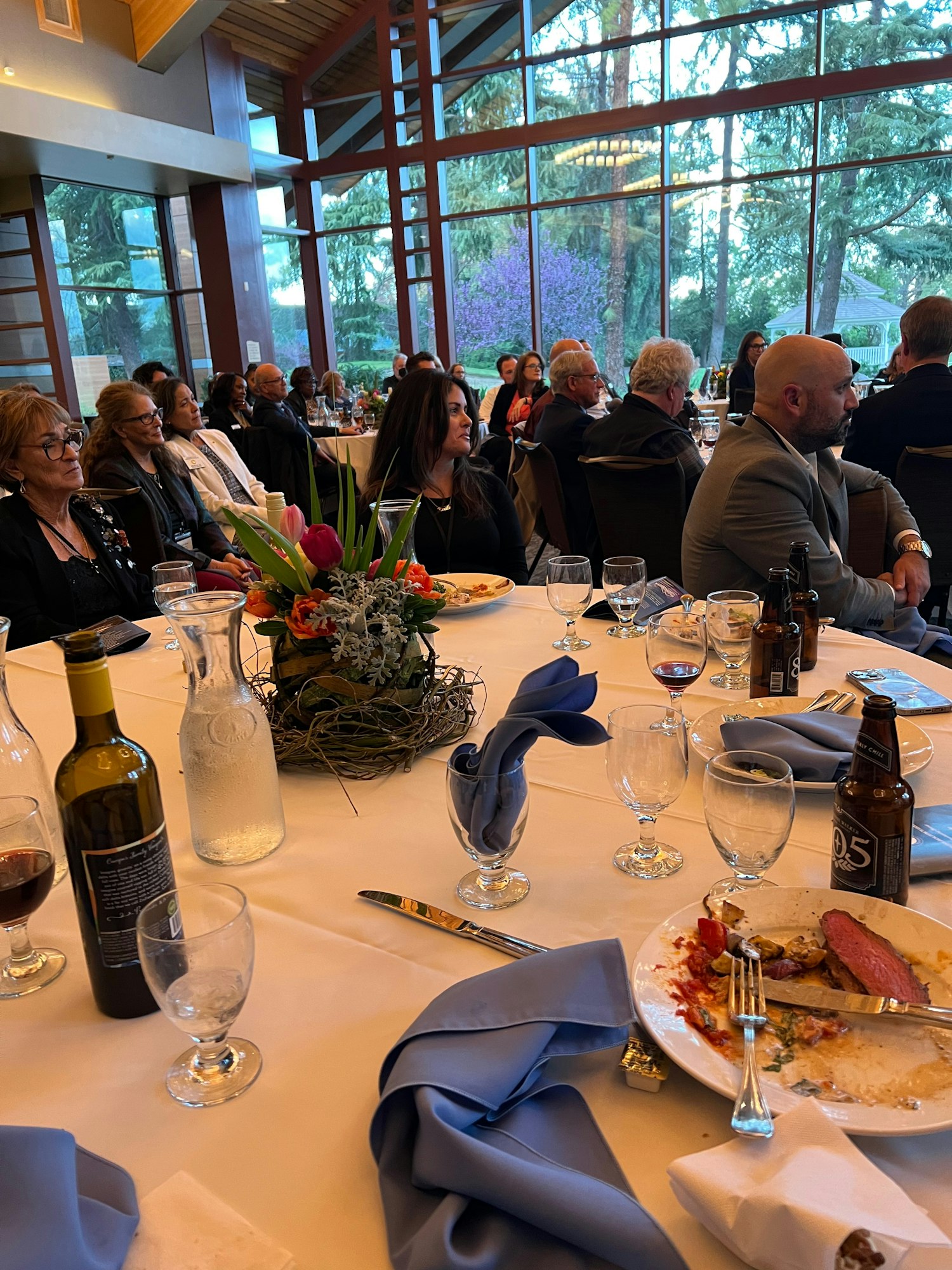 People seated at tables in a event hall with large windows; plates, drinks, and a floral centerpiece on the tables.