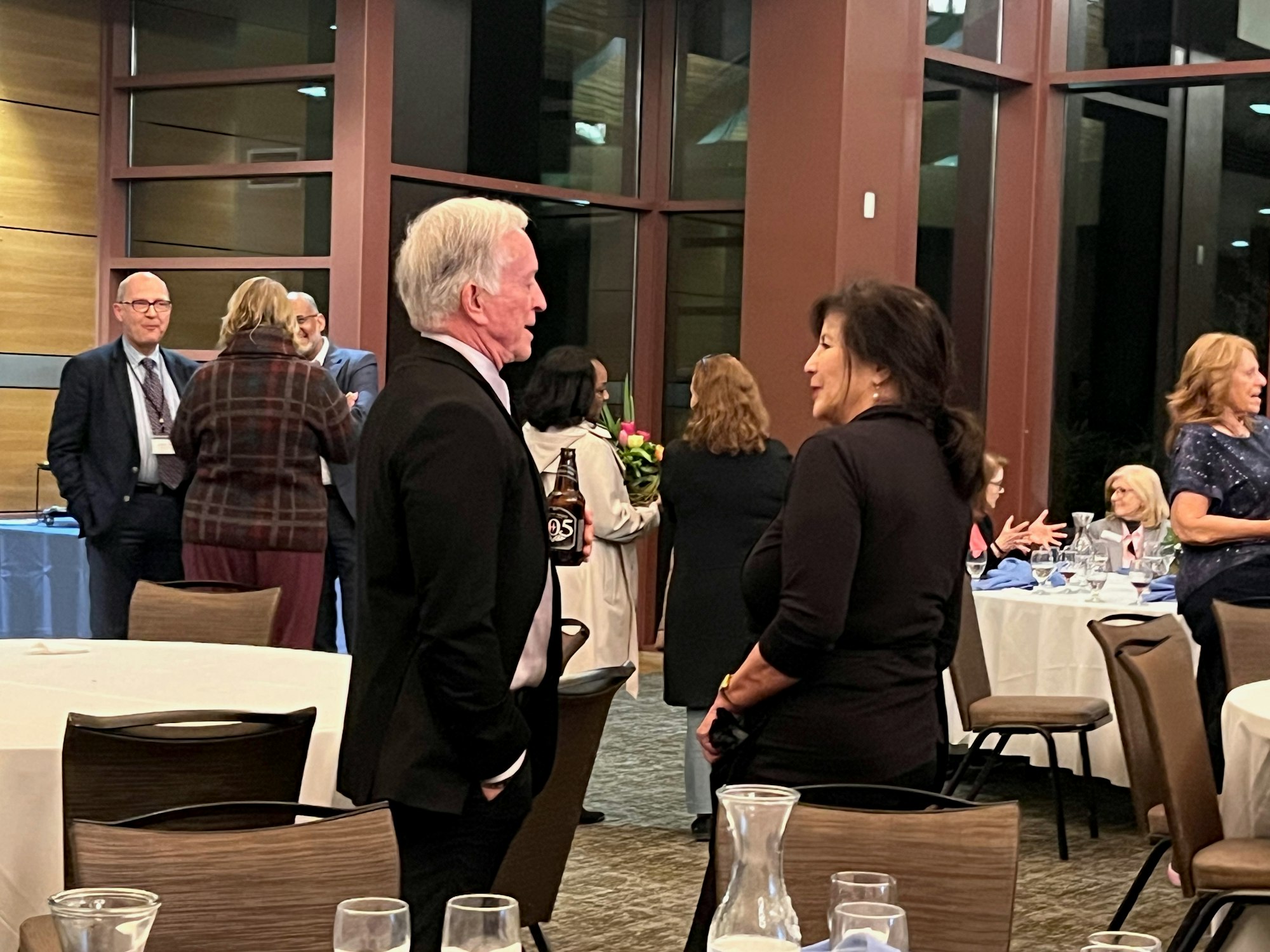 People at a formal event, socializing around tables in a room with large windows.