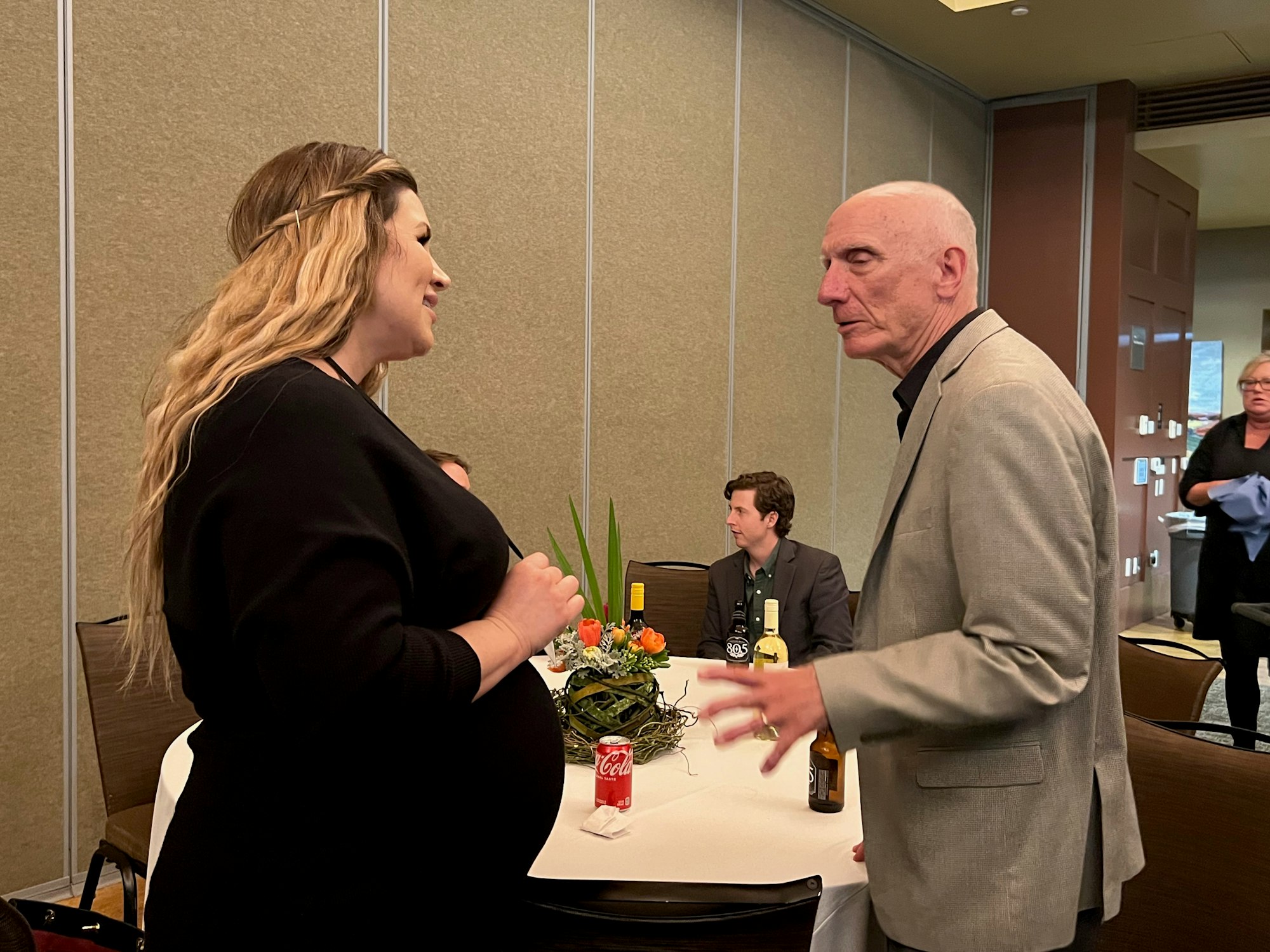Two people are conversing at a table with drinks and a flower arrangement in a conference room setting.