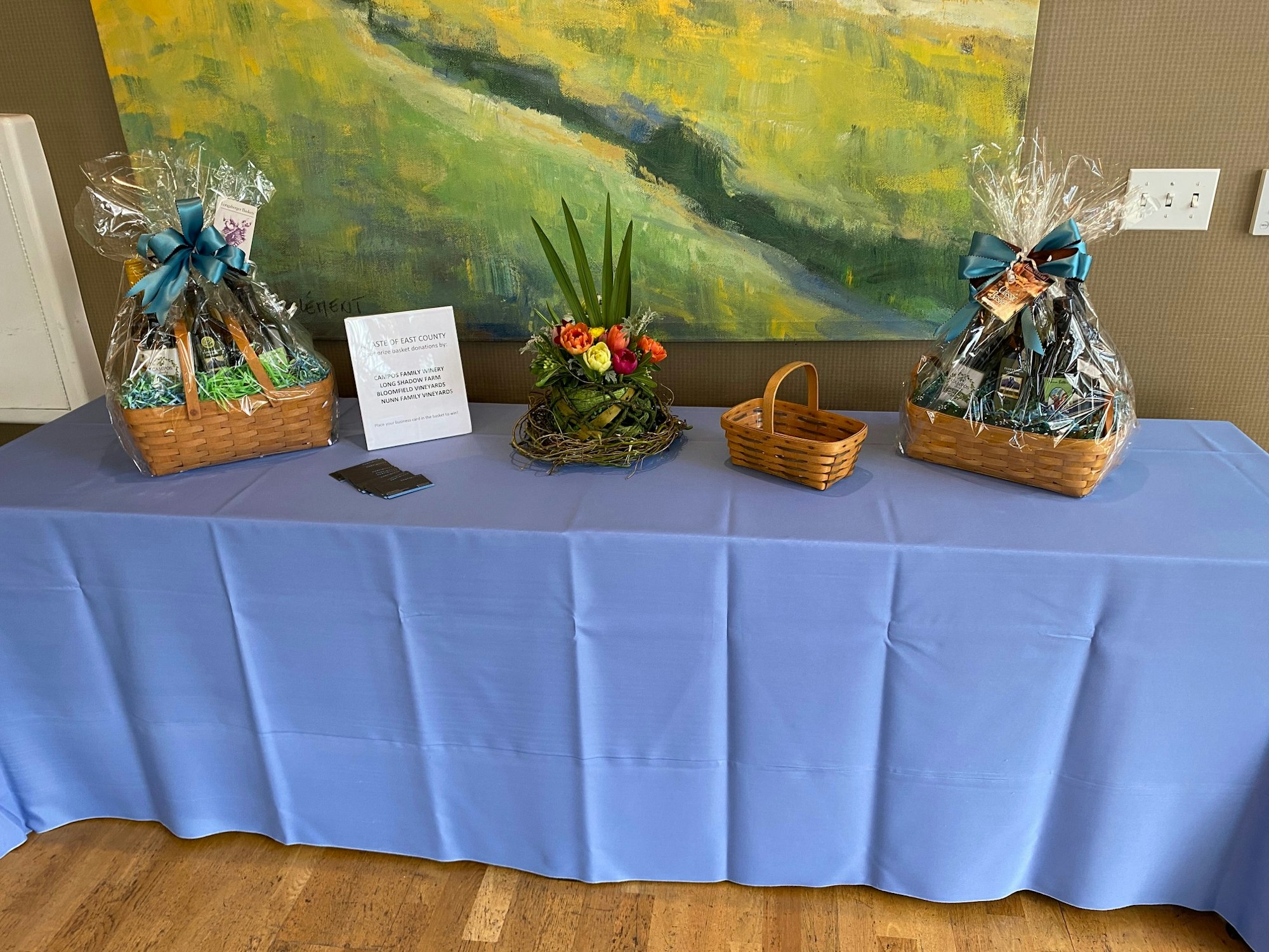 Baskets wrapped in plastic, a flower arrangement, and a sign on a table with a blue cloth.