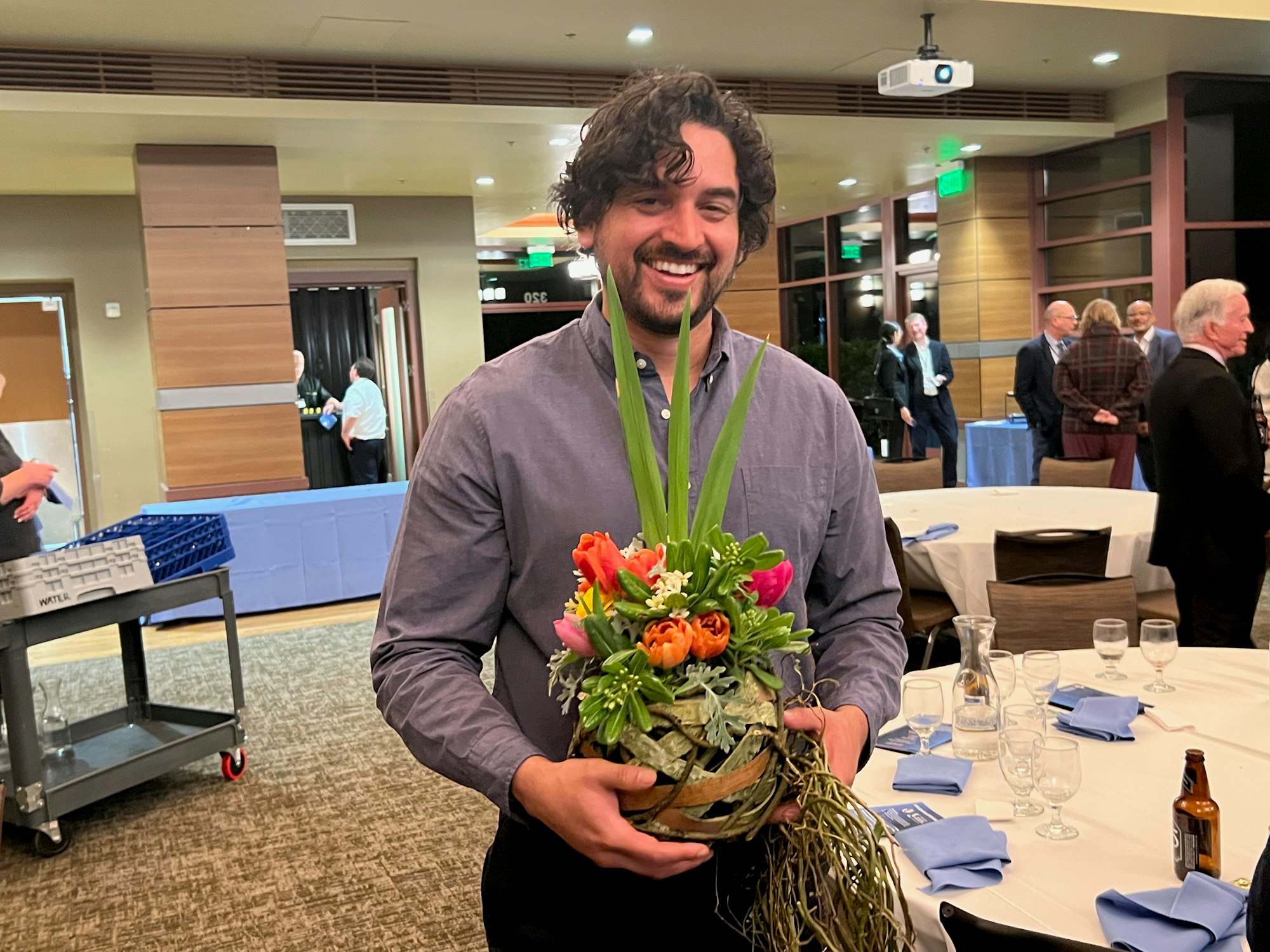 A person smiling while holding a floral arrangement at an event. Guests and tables are visible in the background.