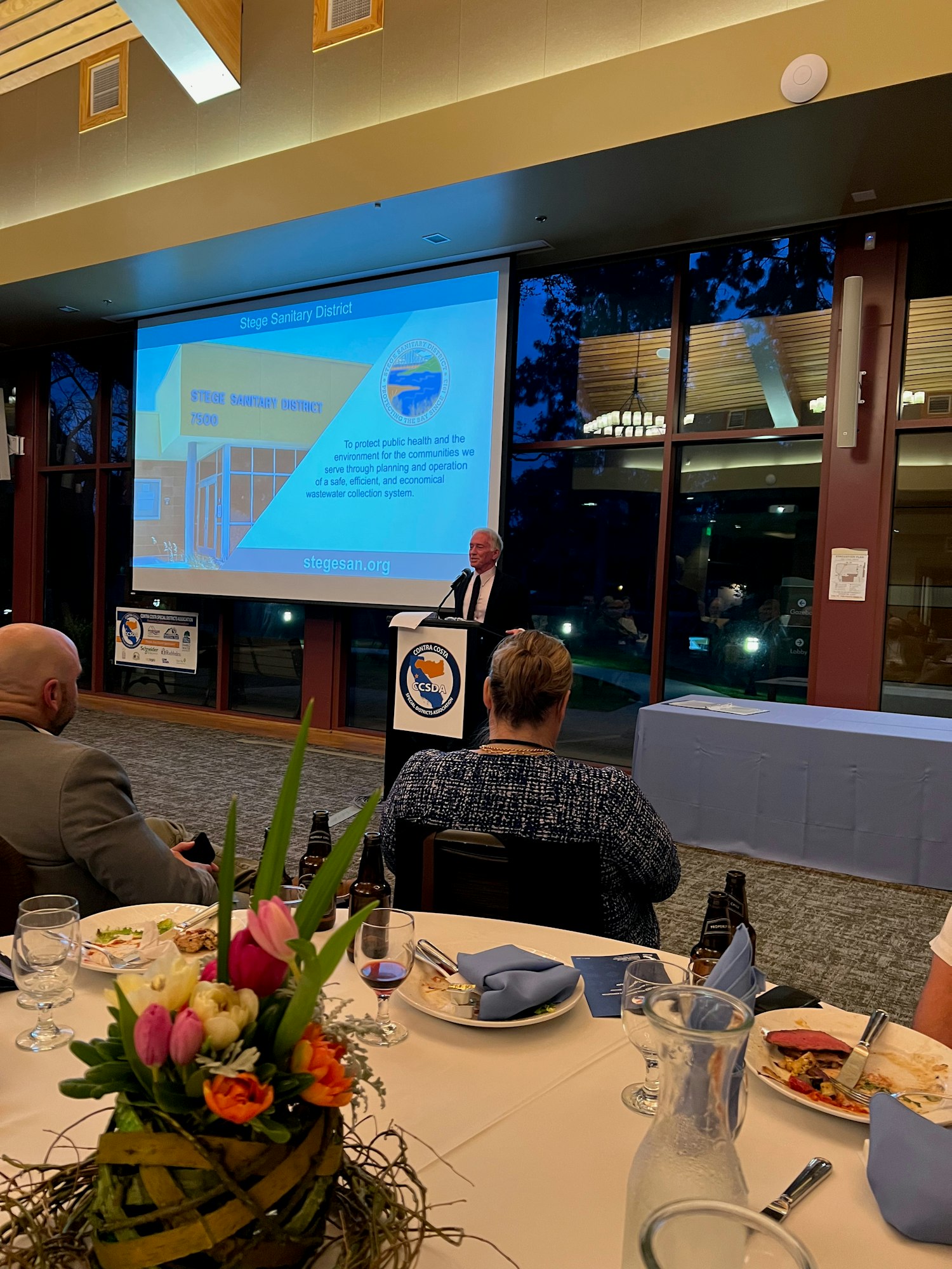 A speaker at a podium presents about the Stege Sanitary District at an event with dinner attendees and floral centerpieces.