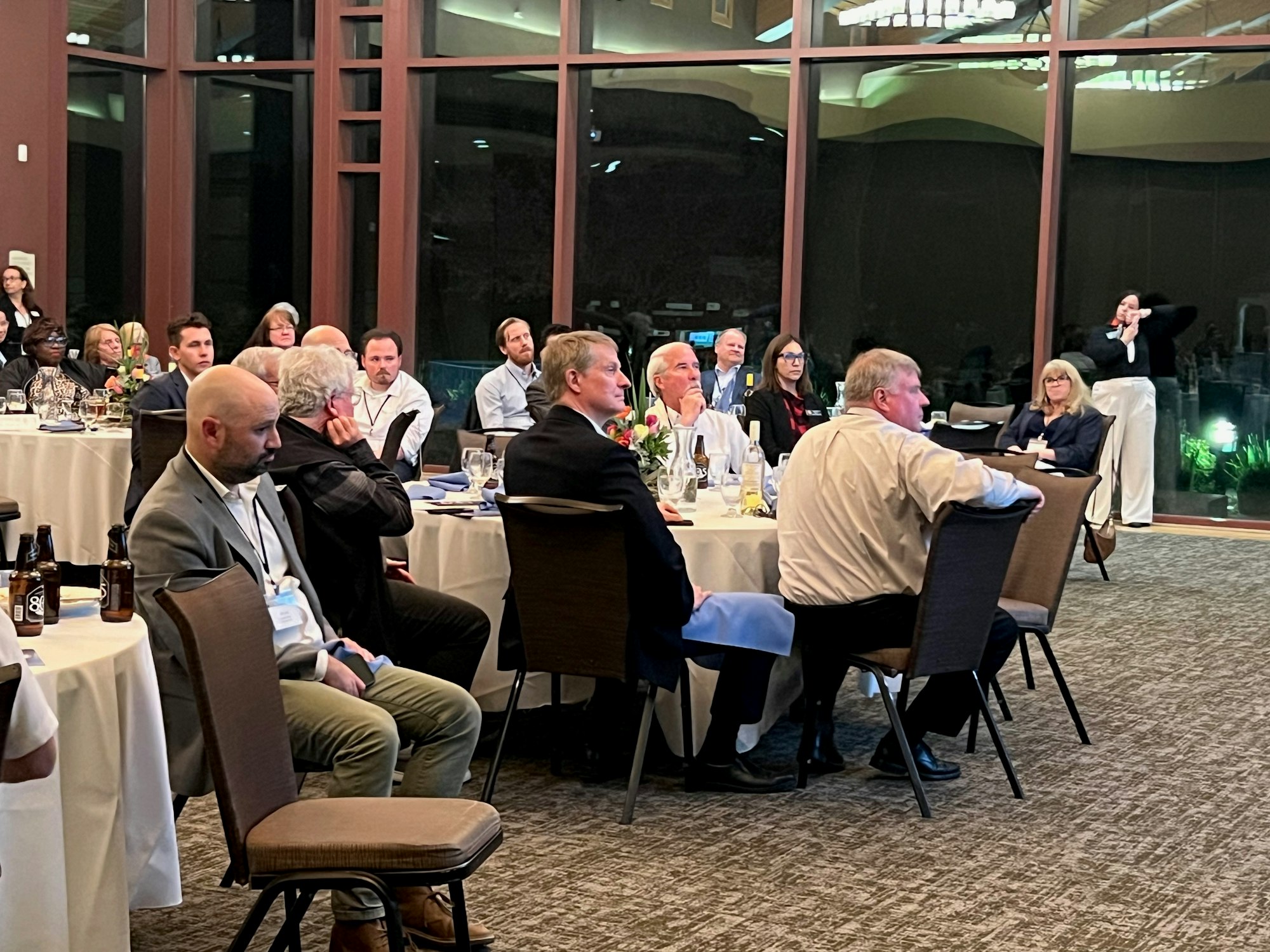 People in formal attire sitting at round tables during an indoor event or presentation.