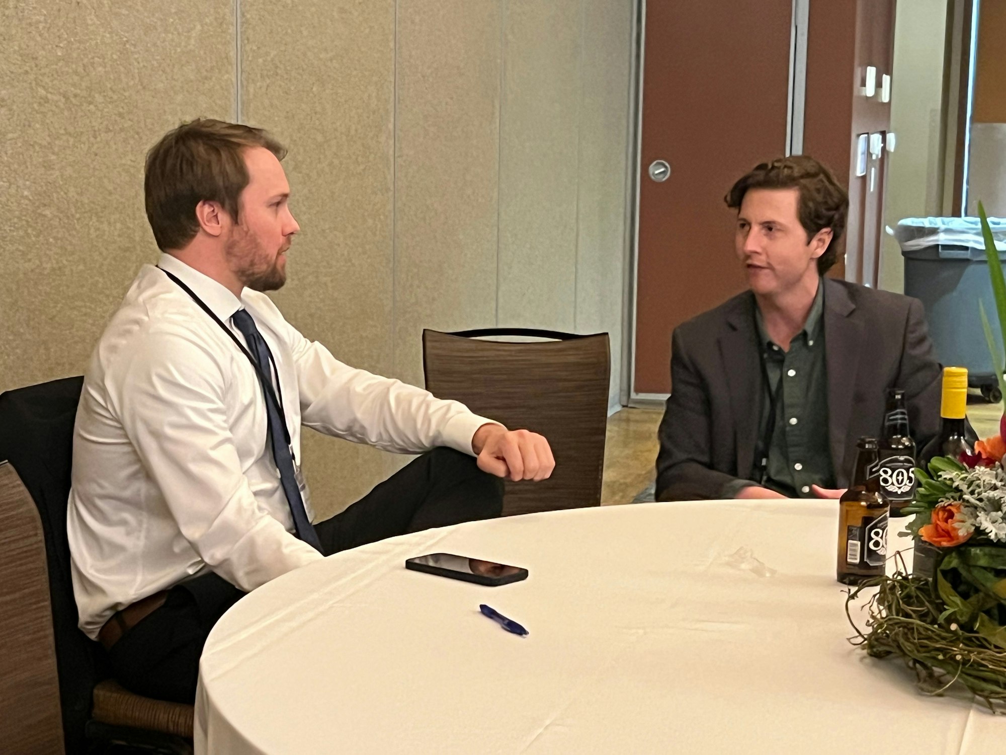 Two men in conversation at a table with drinks and a floral centerpiece.