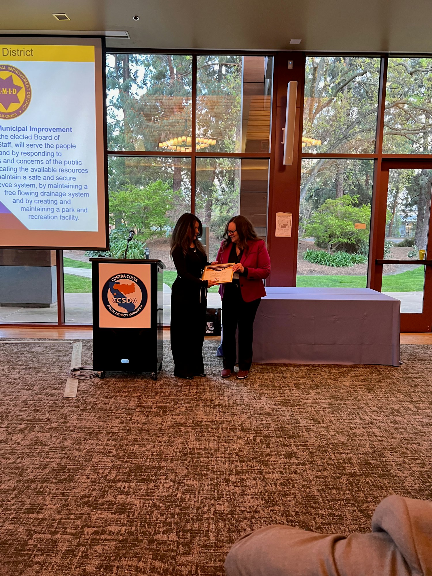 Two people at a ceremony, one receiving a certificate. There's a lectern, a screen with text, and large windows showing greenery.