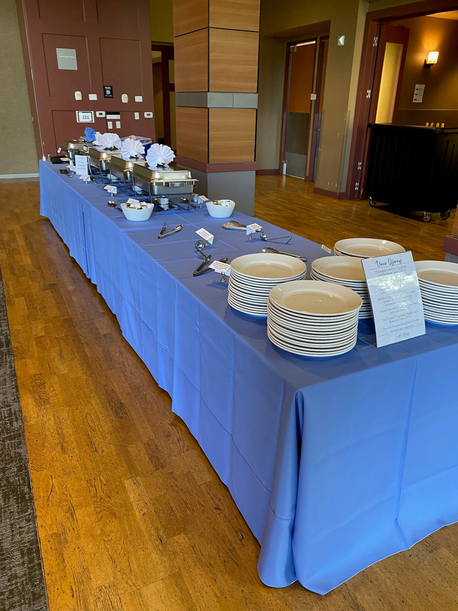 Buffet setup with blue tablecloth, stacked plates, chafing dishes, utensils, and a menu sign.
