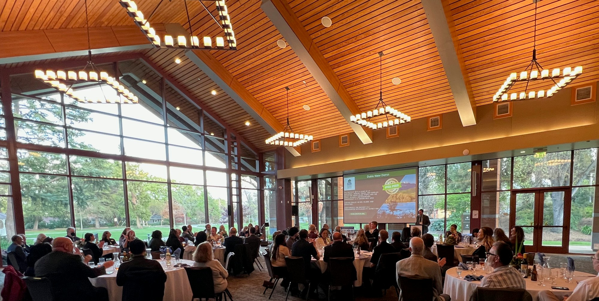 A conference or event with people seated at round tables in a large, well-lit room with high ceilings and a presentation screen.