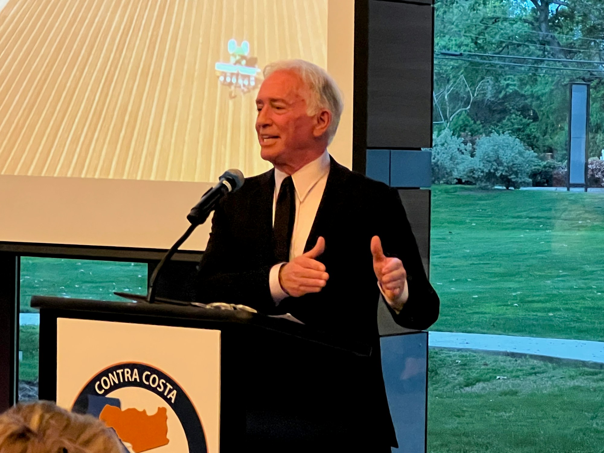 A man in a suit speaking at a podium with a "Contra Costa" emblem, in front of a screen display and window showing a green lawn.