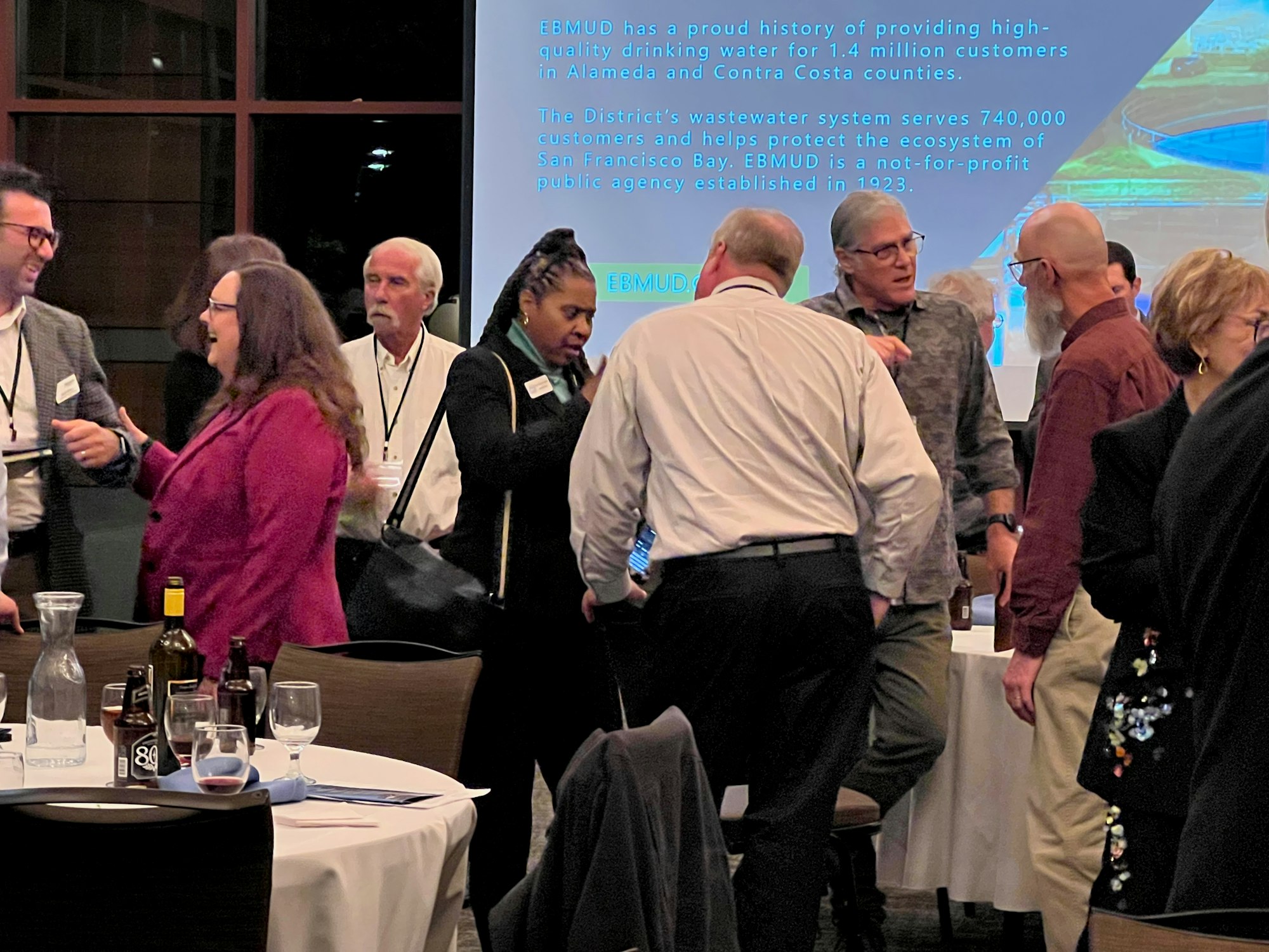 A group of people socializing at an event with a screen in the background displaying text about EBMUD.