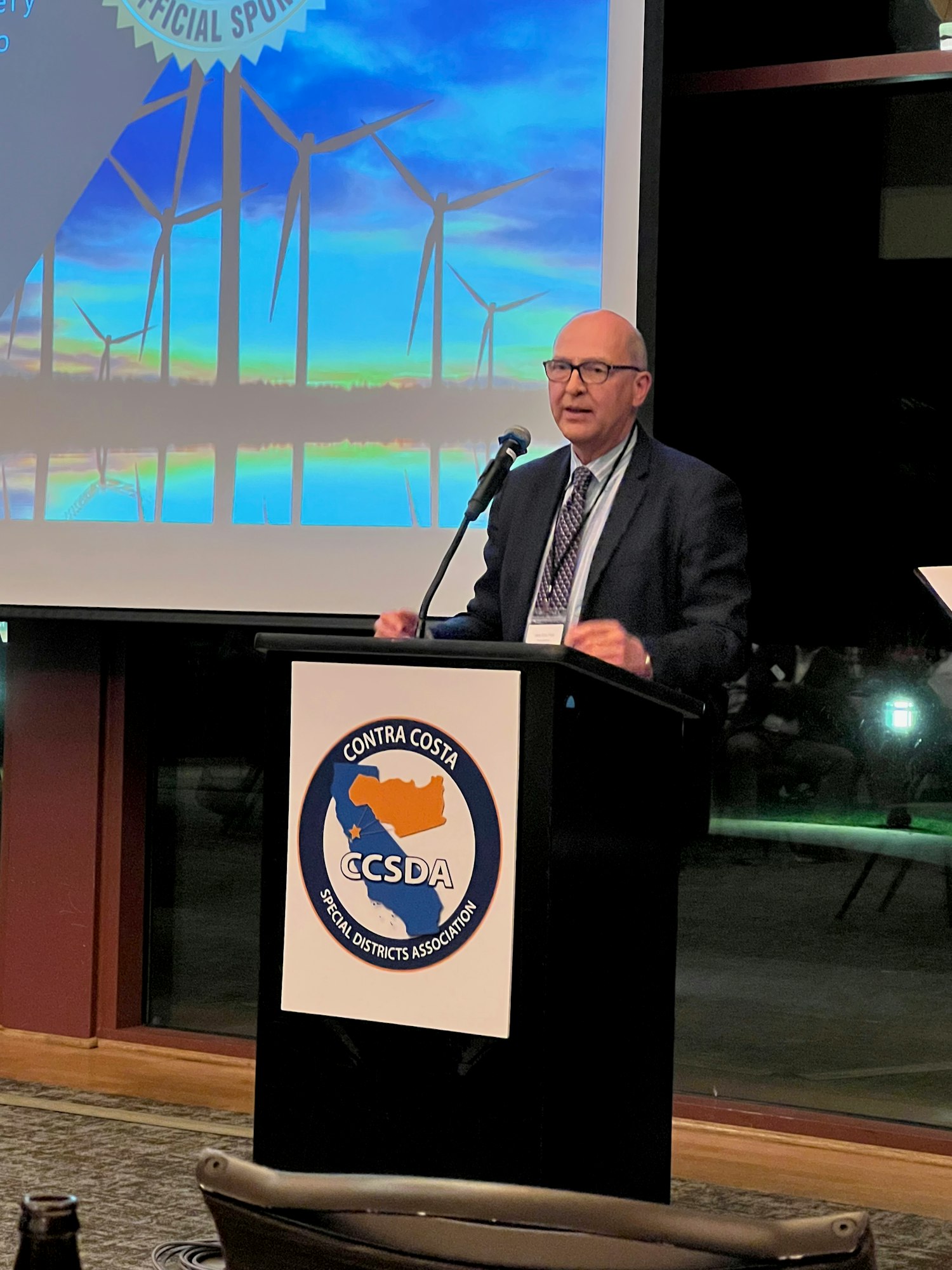 A person speaking at a podium labeled "Contra Costa Special Districts Association" with wind turbines on a screen behind.