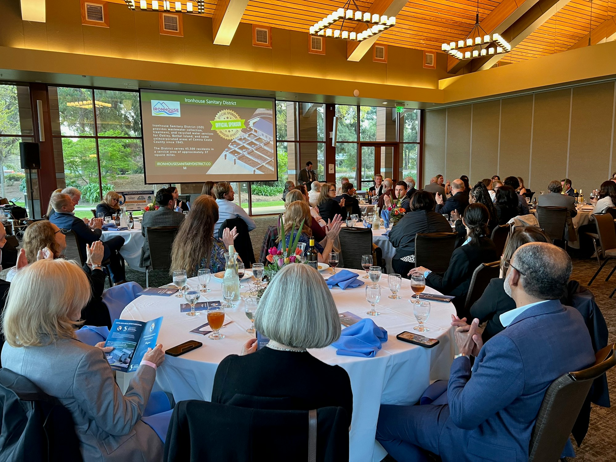 A group of people seated at tables in a banquet hall with a presentation displayed on a screen.