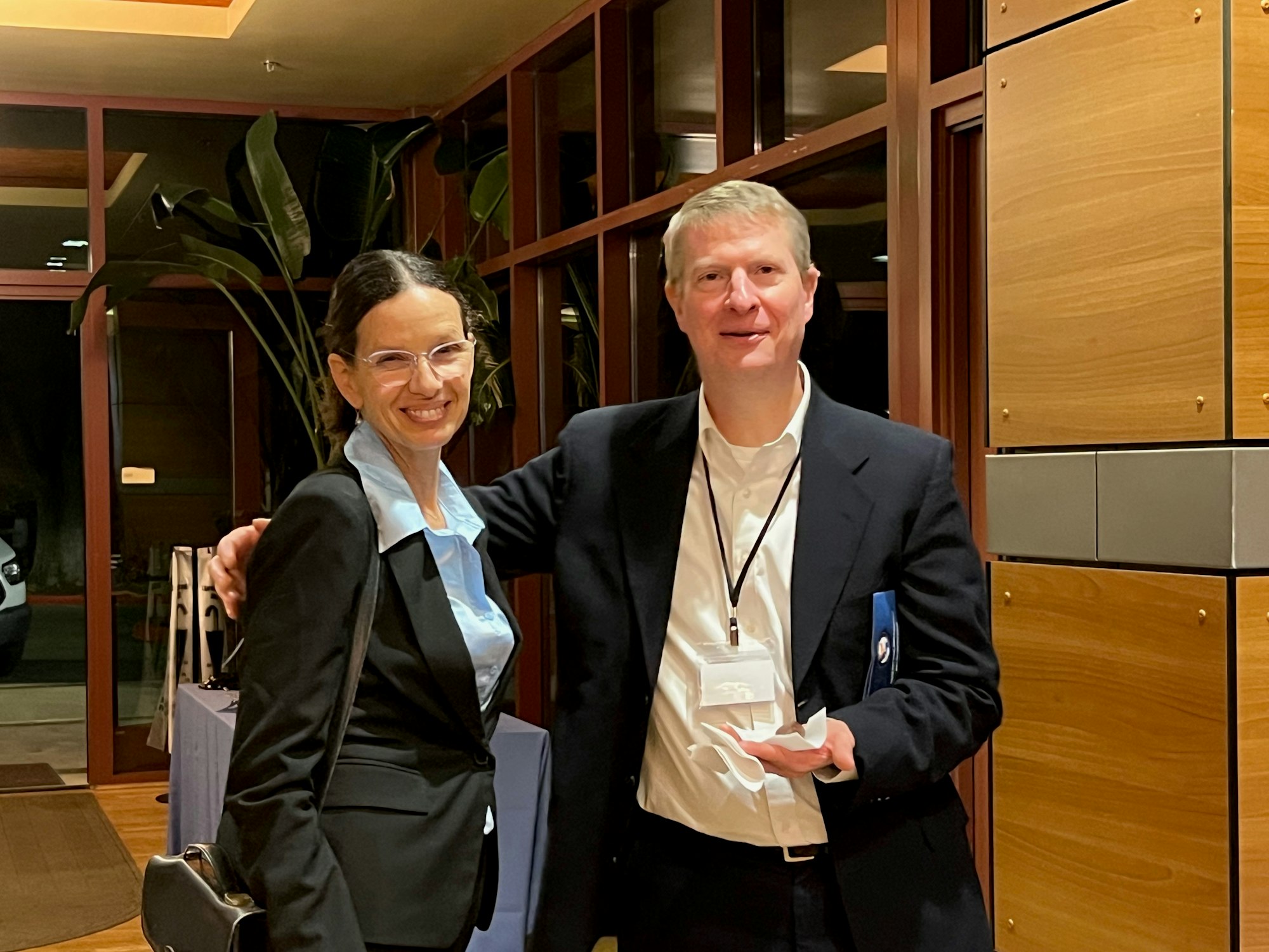 Two people in business attire smiling, standing indoors with wood and glass paneling.