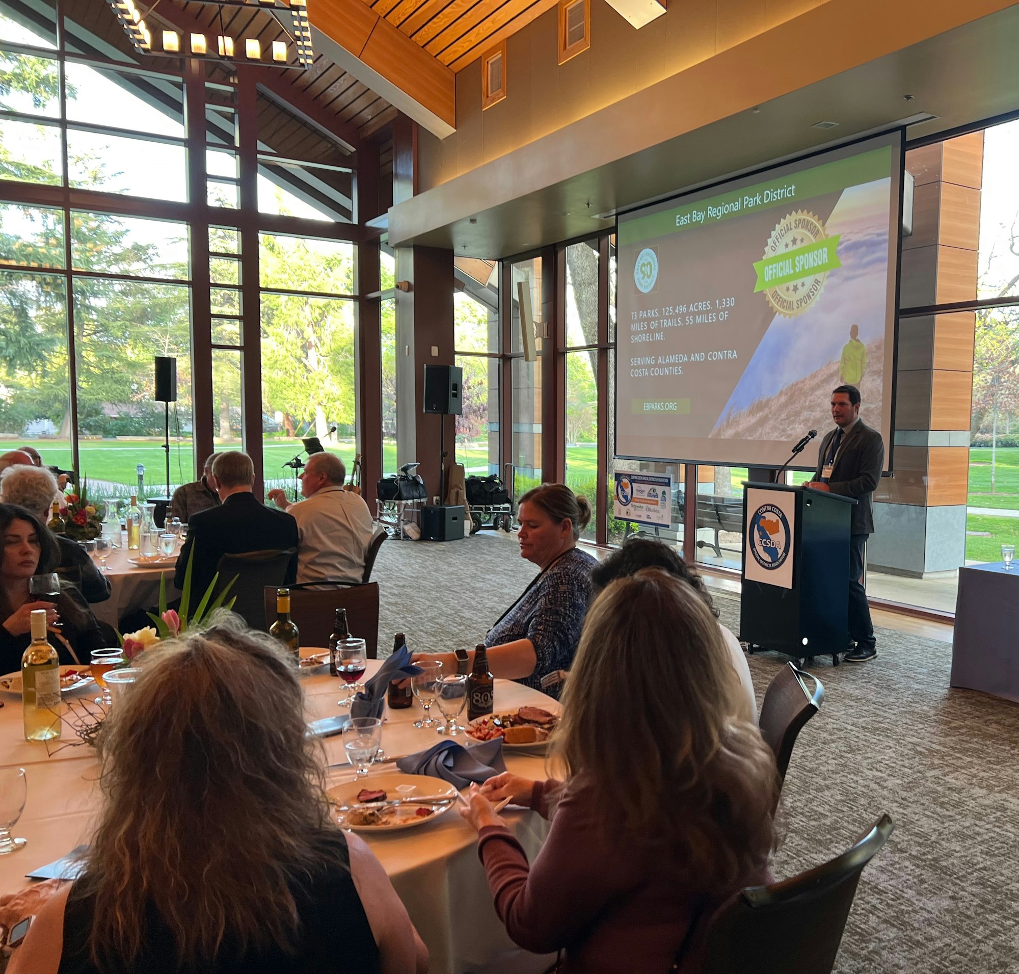 People dining and listening to a presentation at an indoor event with large windows showcasing a garden.