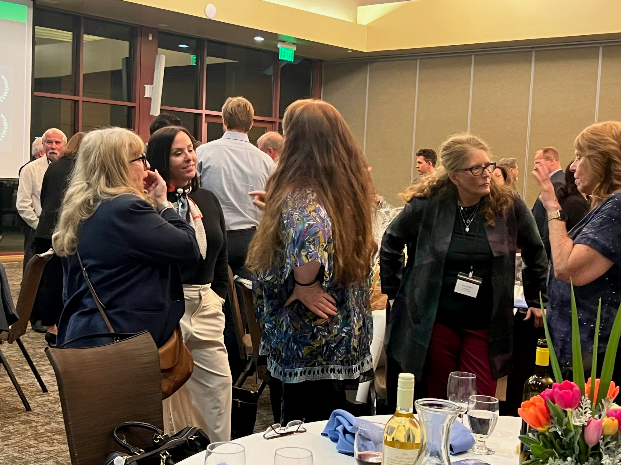 A group of people are talking at an indoor social event with tables, drinks, and flowers.