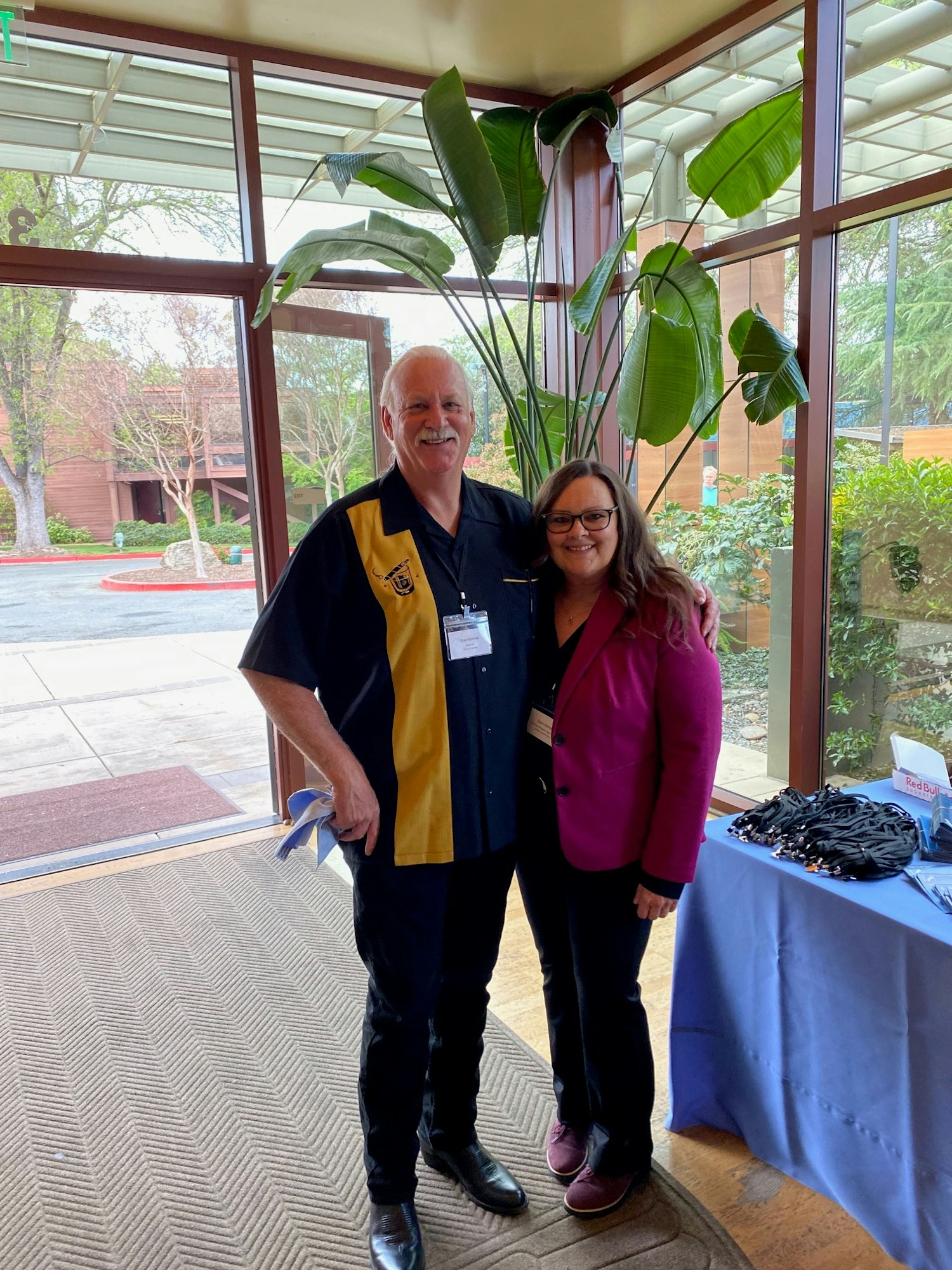 Two people posing indoors near a large plant, with a table and windows in the background.