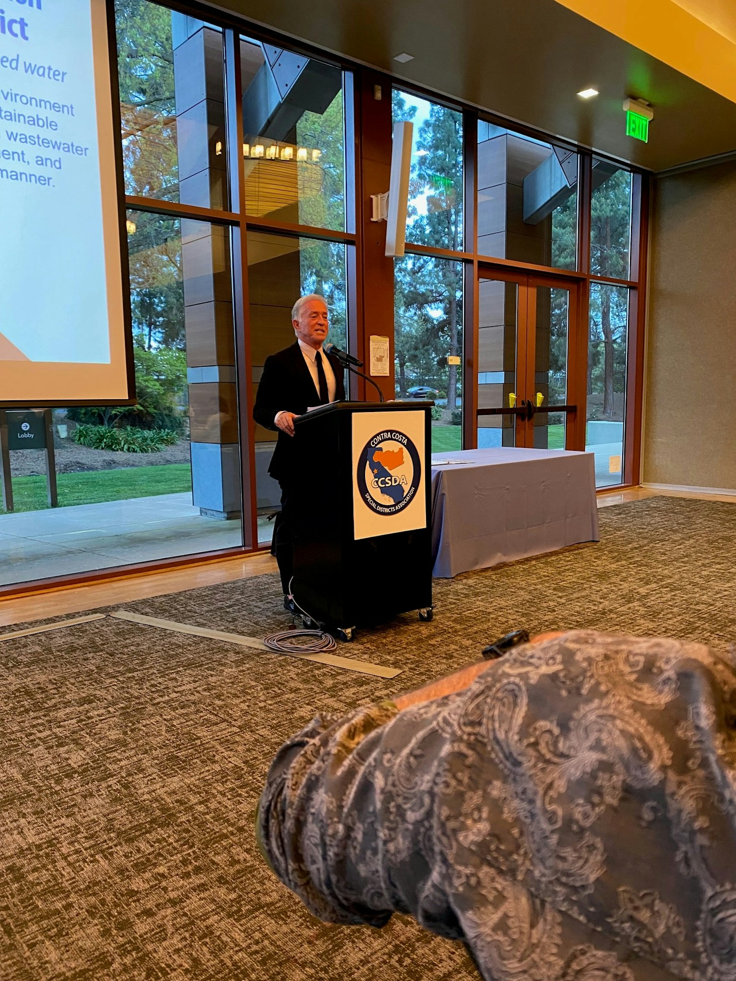 A person speaking at a podium with a CCSDA logo, in a room with large windows and a presentation screen.