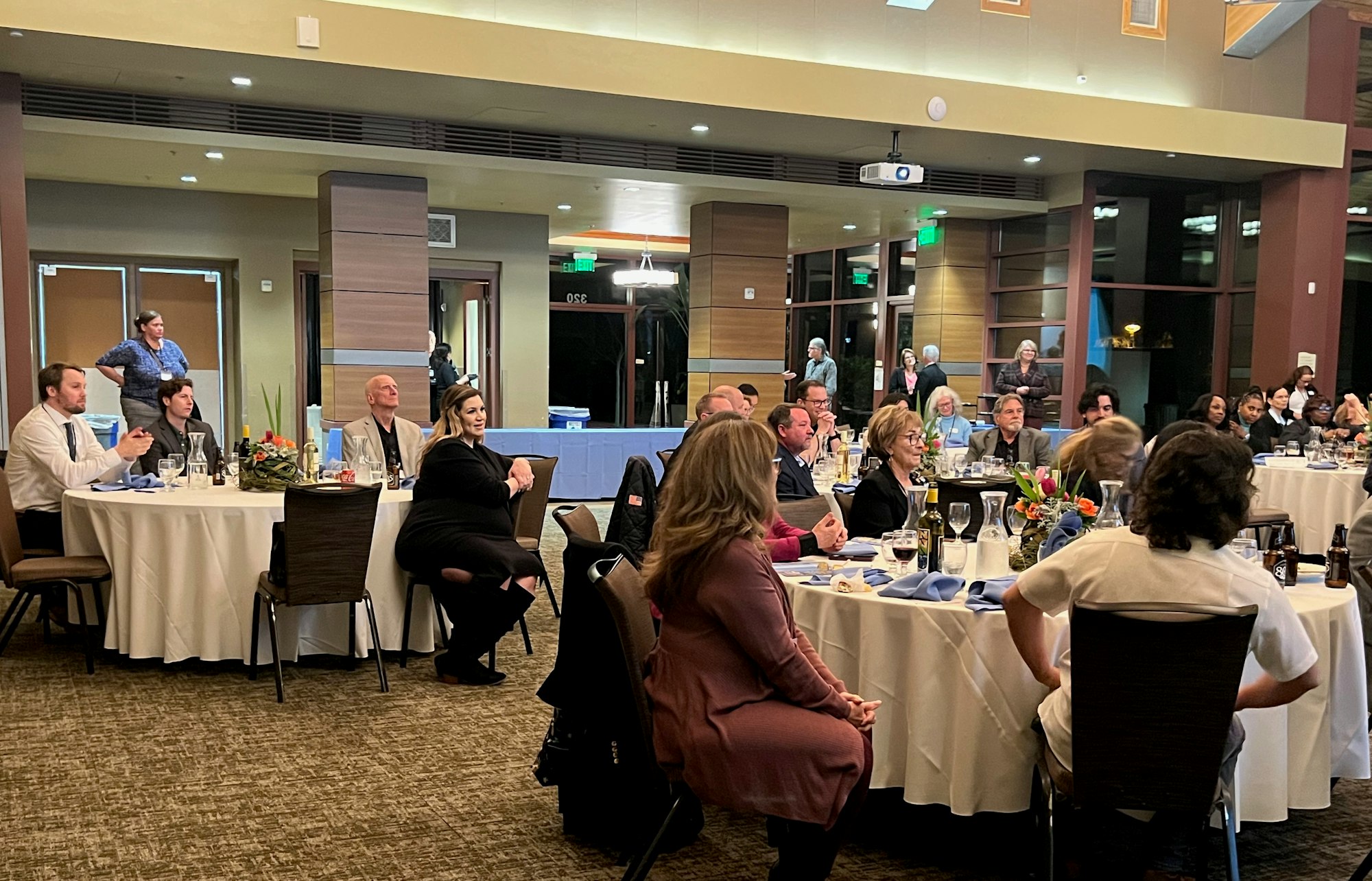 A group of people seated at round tables in a banquet hall, attentively watching something.